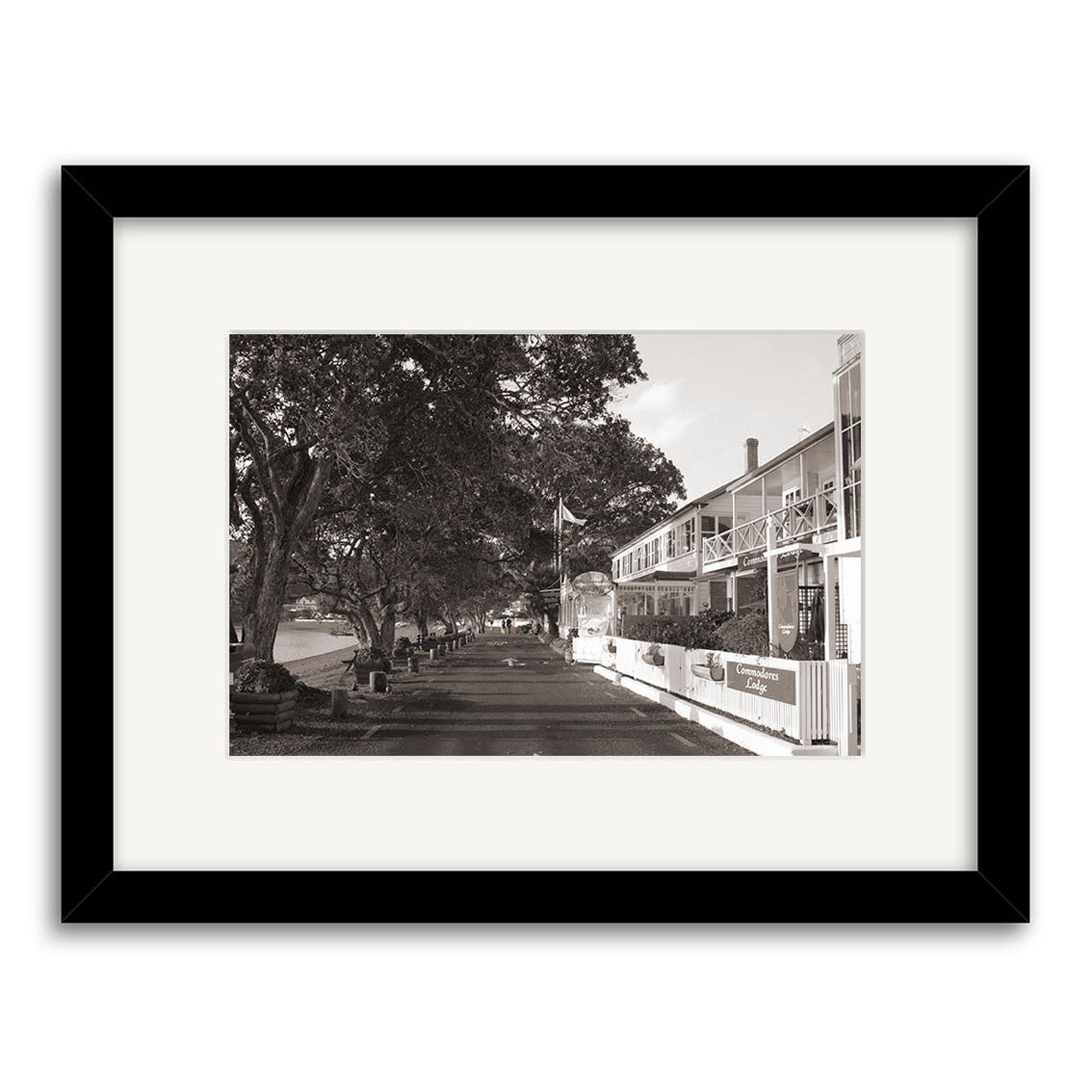 The Strand Russell - RP030 is a black-and-white photo of a tree-lined street with benches and a two-story building, displayed in a black frame with white mat, printed on archival quality matte paper.