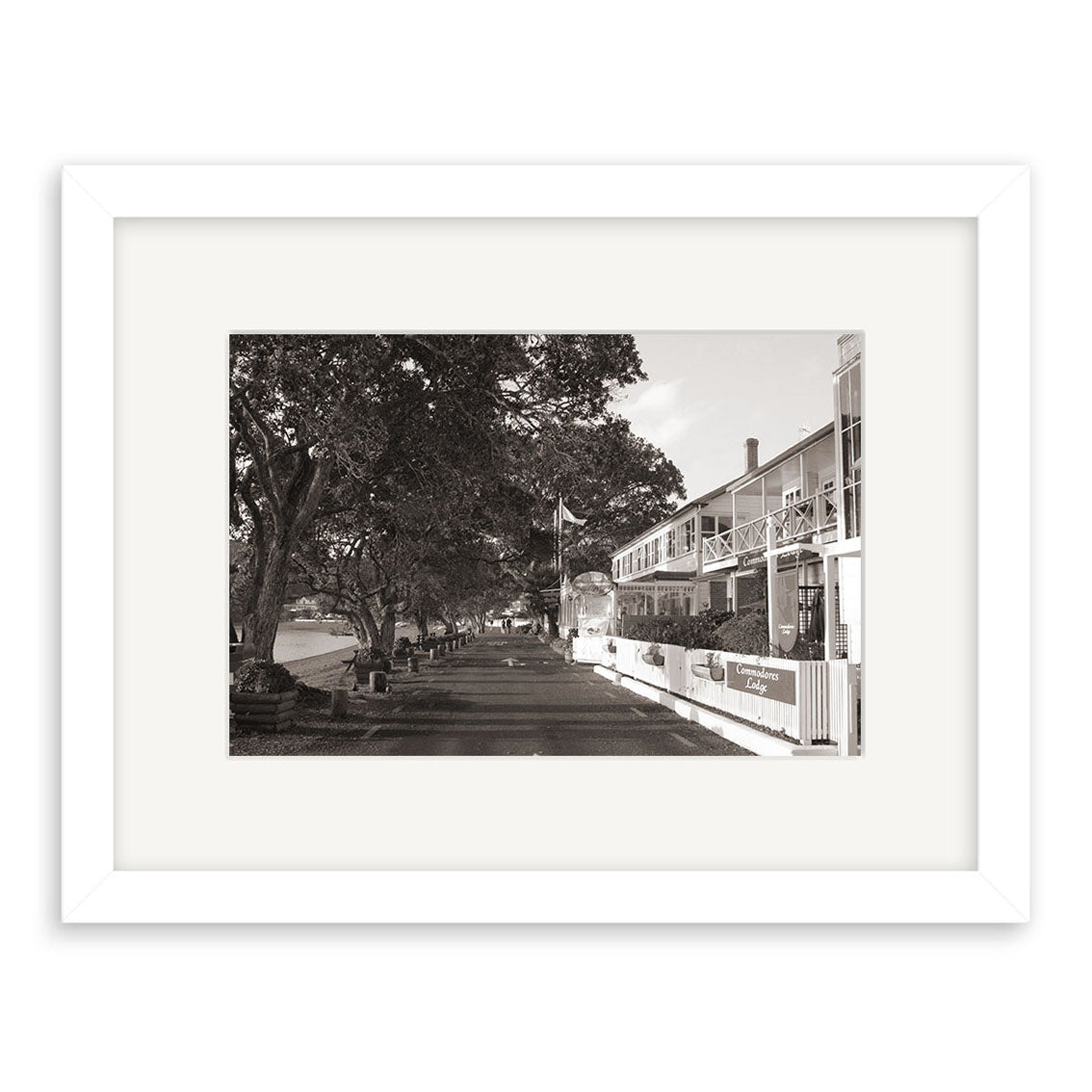 The Strand Russell - RP030: Black and white photo of a tranquil New Zealand street with trees, a two-story building, balconies, and a café. Presented in a white frame—ideal as an archival quality matte photographic print.