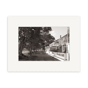 The Strand Russell - RP030 is a black and white photo of a tree-lined street in New Zealand, benches on the left, and a two-story building with balcony and signs on the right. Available as photographic prints on archival matte paper.