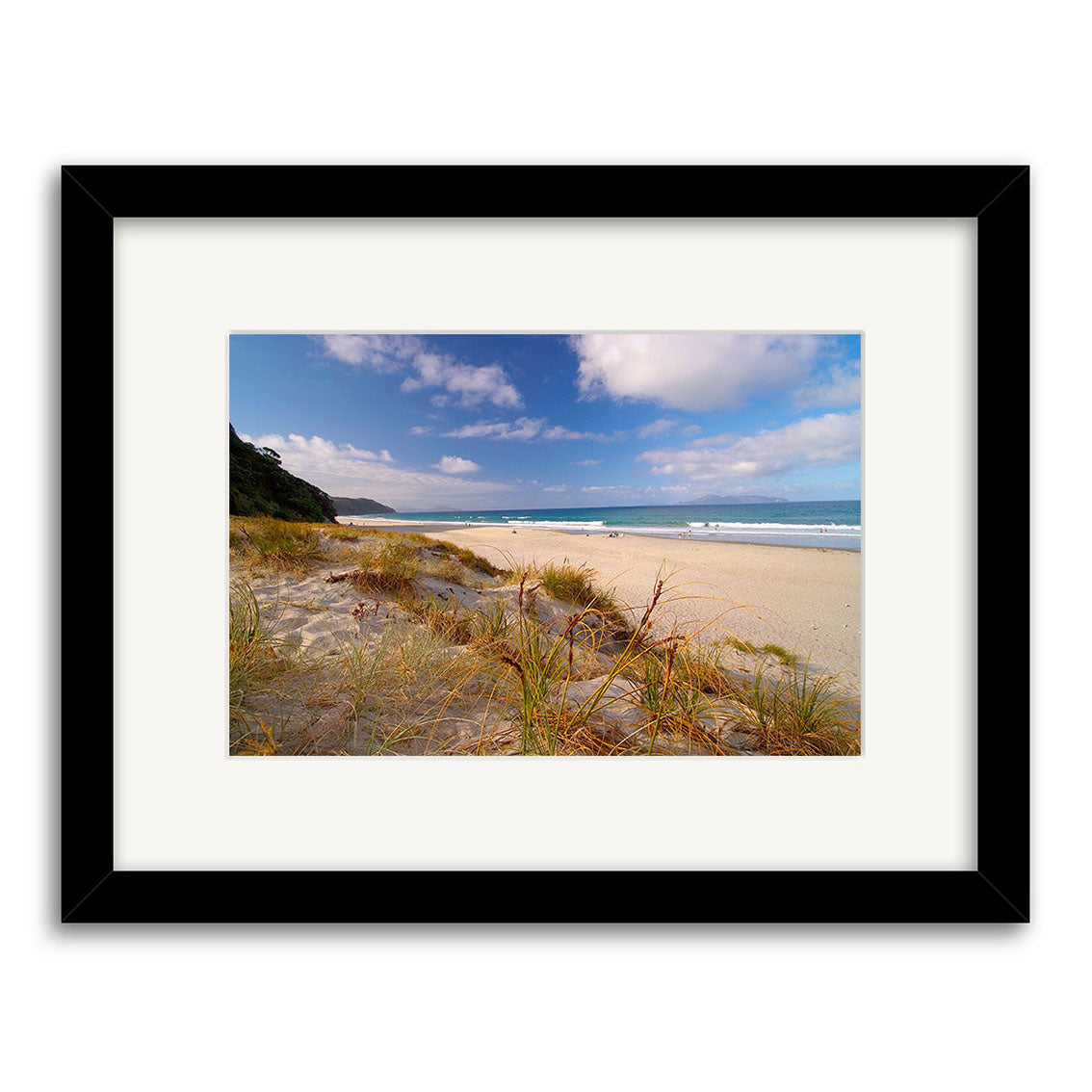 Mangawhai Beach - RP032: Framed archival print featuring a sandy New Zealand beach with grassy foreground, blue waves, and a partly cloudy sky. Presented centered in a white mat within a black frame.
