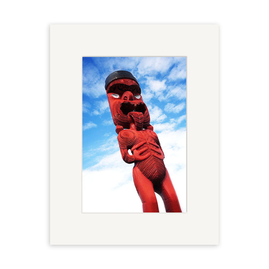 The Maori Statue - RP041 is a red wooden Maori figure with large eyes and intricate patterns, photographed against a blue sky and framed with a white border—available as high-quality photographic prints on archival paper.