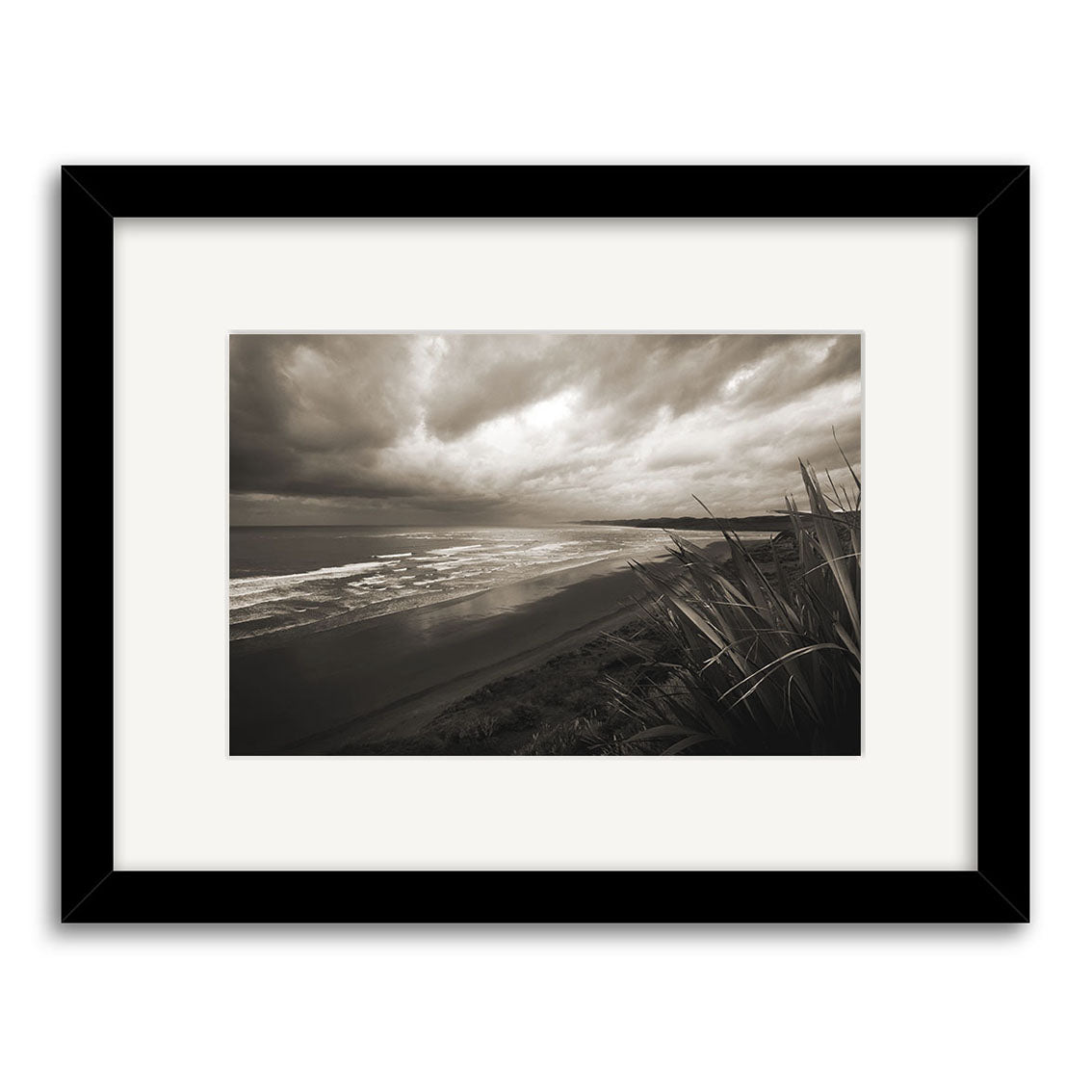 Raglan - RP042: Black and white framed photograph of a New Zealand coastal landscape, featuring cloudy skies, ocean waves, sandy shore, and tall grass. Expertly printed on archival quality matte paper.