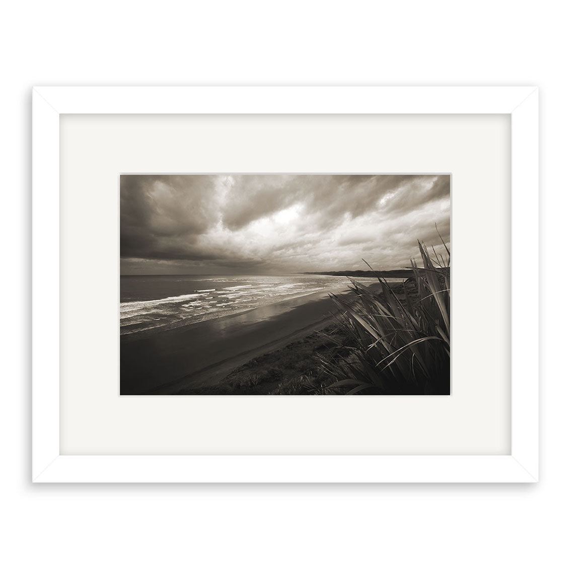 Raglan - RP042 is a black and white photo of a New Zealand beach with waves and dark clouds, framed by tall grass on the right. Displayed in a white mat and frame, this archival quality matte print adds elegance to any space.