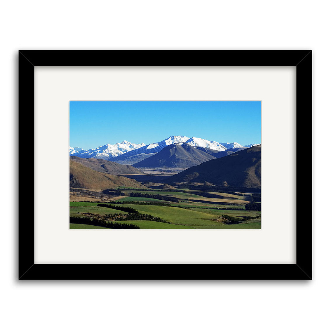 Inverary - RP046 is a framed photo print of green fields, rolling hills, and snow-capped mountains under a blue sky. Printed on archival matte paper, it includes a white mat and sleek black frame for a classic finish.