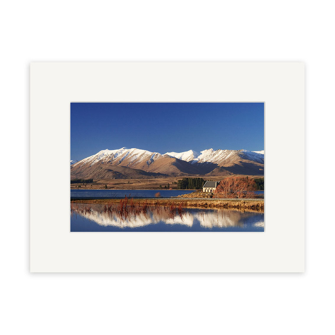 A small house stands by Lake Tekapo in New Zealand, snow-capped peaks behind it and dried grasses reflected in the calm water—a stunning scene for photographic prints on archival quality matte paper. Product: Lake Tekapo - RP047.