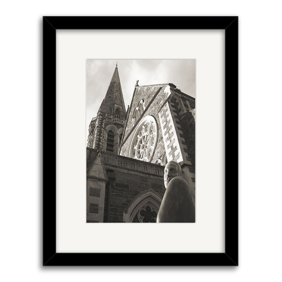 Christchurch Cathedral - RP048 is a black and white framed photographic print by Reuben Price, featuring a stone church with a tall steeple and stained glass window in New Zealand, with a statue thoughtfully gazing in the foreground.