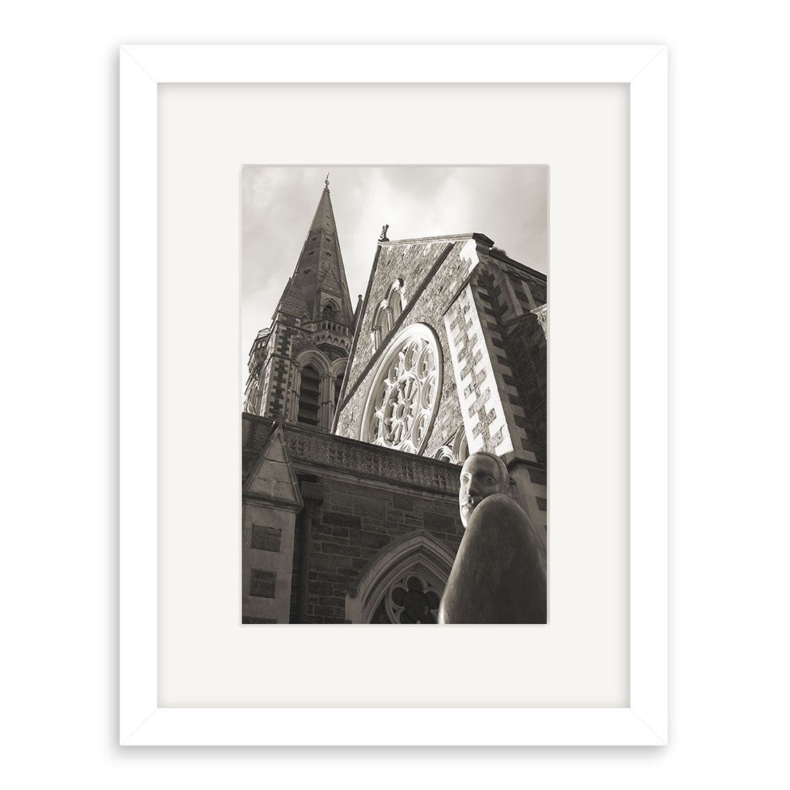 Black and white photo of Christchurch Cathedral - RP048, featuring a stone church with tall spire, pointed arch windows, and rose window. A statue stands in front. Photographic print by Reuben Price, framed in white.