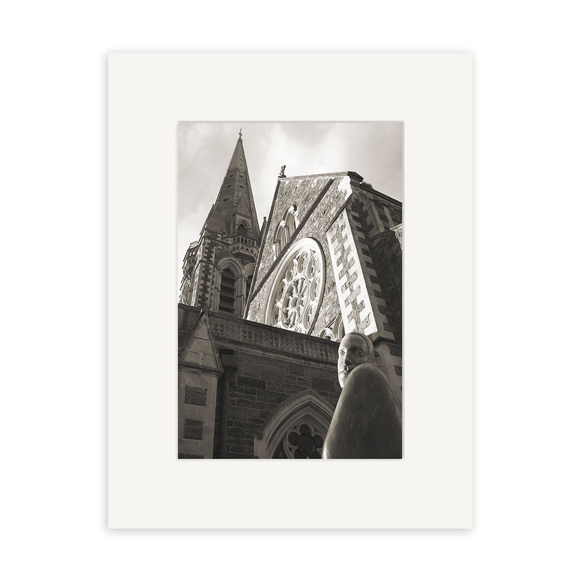 Christchurch Cathedral - RP048: a black and white photo of a stone church with tall spire, circular window, and foreground statue by Reuben Price, printed and centered on a plain white mat.
