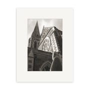 Christchurch Cathedral - RP048: a black and white photo of a stone church with tall spire, circular window, and foreground statue by Reuben Price, printed and centered on a plain white mat.