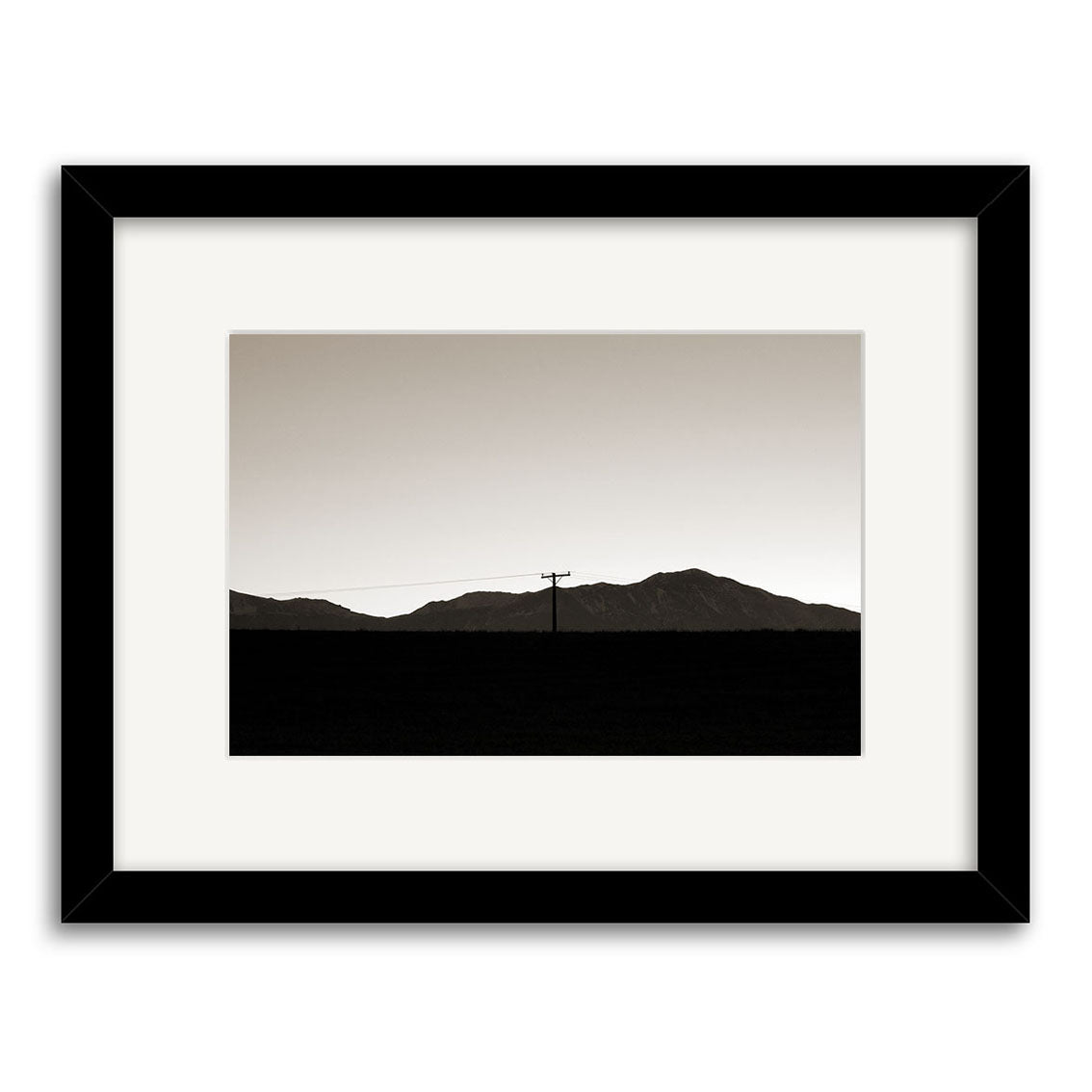 Canterbury Dusk - RP050: An archival-quality black and white photograph of a distant power line before low New Zealand mountains under a clear sky, presented with a white mat and simple black frame.