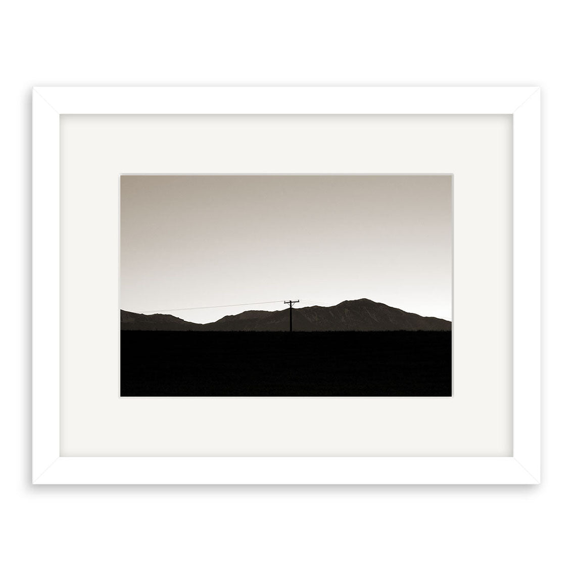Canterbury Dusk - RP050 is a framed black-and-white photo print of a silhouetted New Zealand mountain range and utility pole under a clear sky, presented with archival quality, a wide white mat, and a simple white frame.