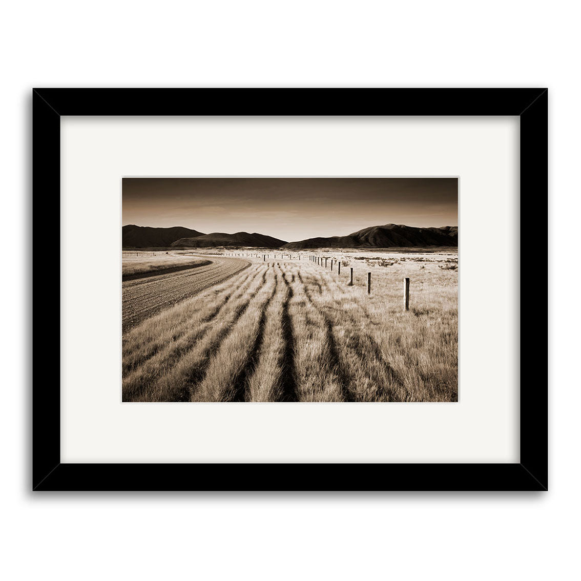 Canterbury back Country - RP052 is a framed black-and-white photograph by Reuben Price, showcasing a rural New Zealand landscape with a curving dirt road, fence line, open fields, and distant mountains—ideal for fans of photographic prints.
