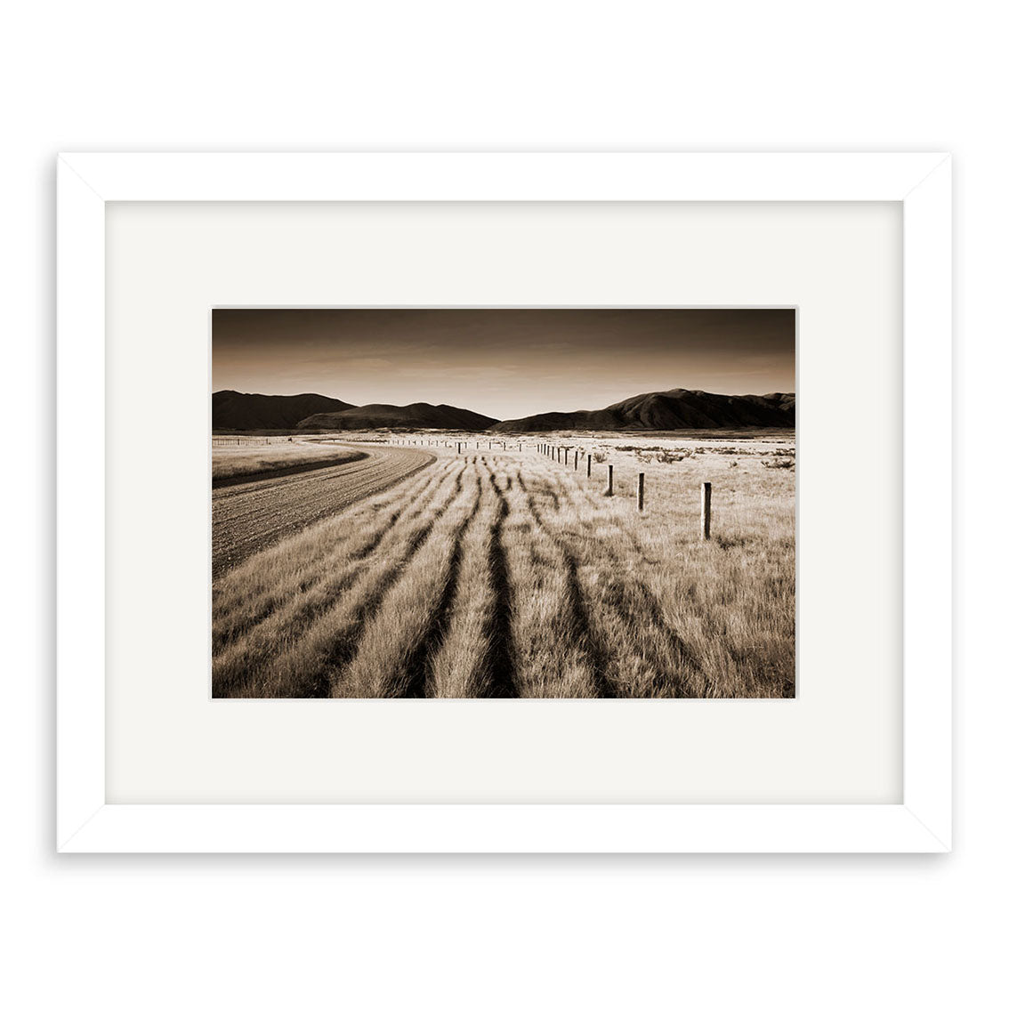 The framed print Canterbury back Country - RP052 by Reuben Price depicts rural New Zealand: tall grass, a curving dirt road, and distant mountains under clear sky in sepia tones.