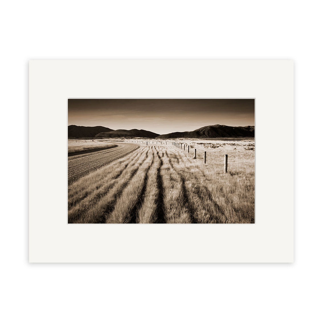 Canterbury back Country - RP052 is a sepia-toned Reuben Price photographic print depicting a rural New Zealand landscape with a dirt road, fence posts, tall grass, and distant mountains, presented with a white mat border.