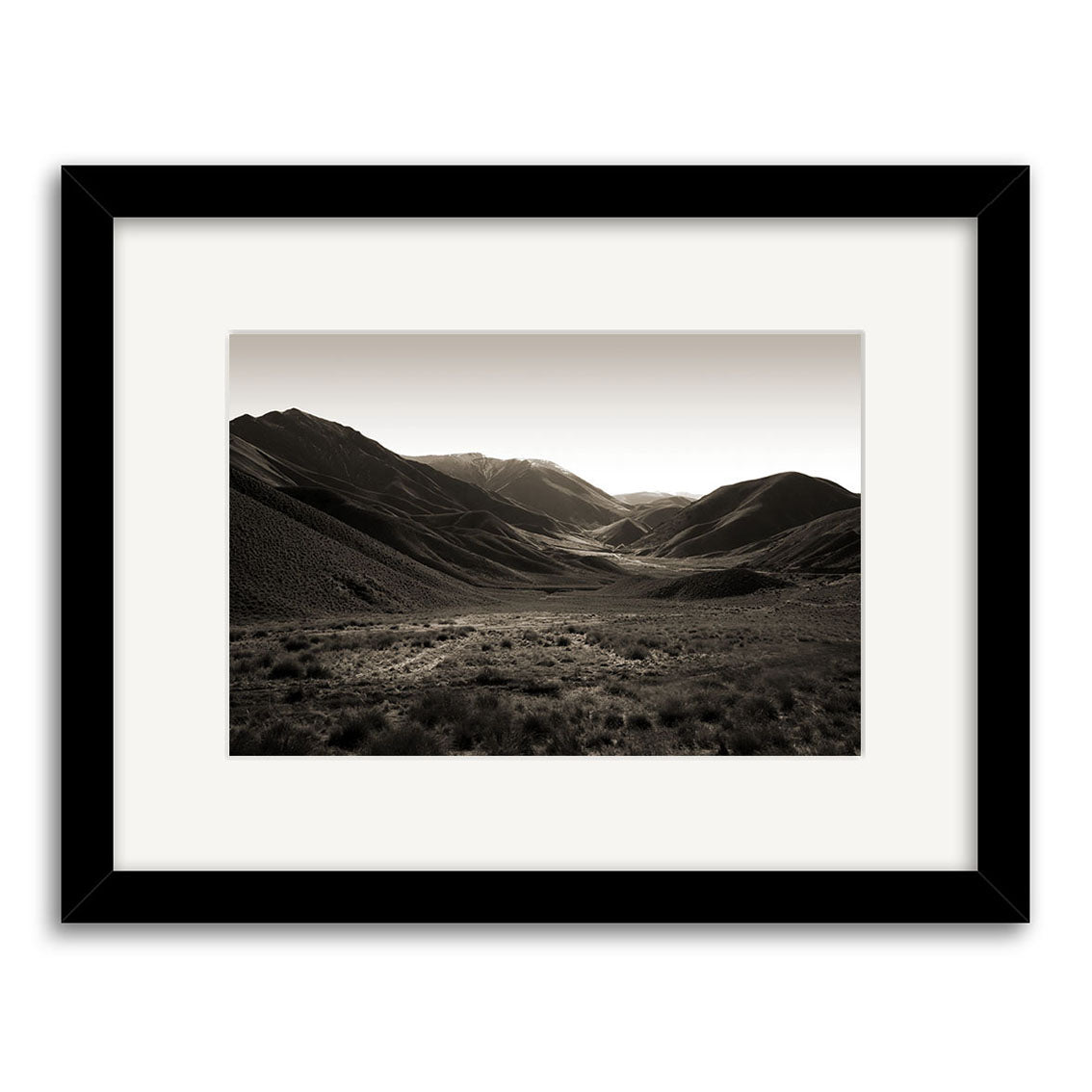 Lindis Pass - RP053 is a framed black-and-white photo of a wide New Zealand valley with rolling hills and distant mountains under a clear sky. Printed on archival matte paper, it includes sparse foreground vegetation and a black frame with white mat.