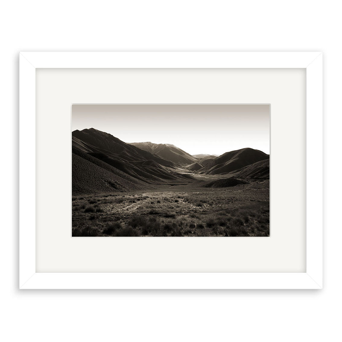 Lindis Pass - RP053: Black and white photographic print of a wide New Zealand valley with dark hills and distant sunlight, framed in white with a mat, printed on archival quality matte paper for lasting beauty.