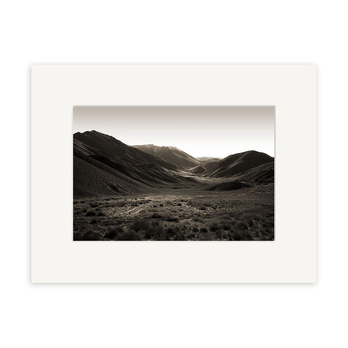 “Lindis Pass - RP053” is a black and white photograph of a wide New Zealand valley ringed by hills and mountains, with grasses and shrubs in the foreground, presented with a white mat border on archival matte paper.