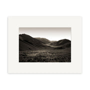 “Lindis Pass - RP053” is a black and white photograph of a wide New Zealand valley ringed by hills and mountains, with grasses and shrubs in the foreground, presented with a white mat border on archival matte paper.