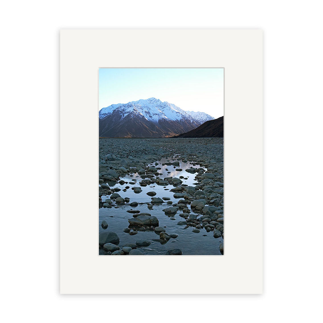 Rangitata River - RP054 features a photo of a rocky riverbed with shallow water reflecting a snow-capped mountain in New Zealand, framed with a simple white mat and printed on archival quality matte paper.