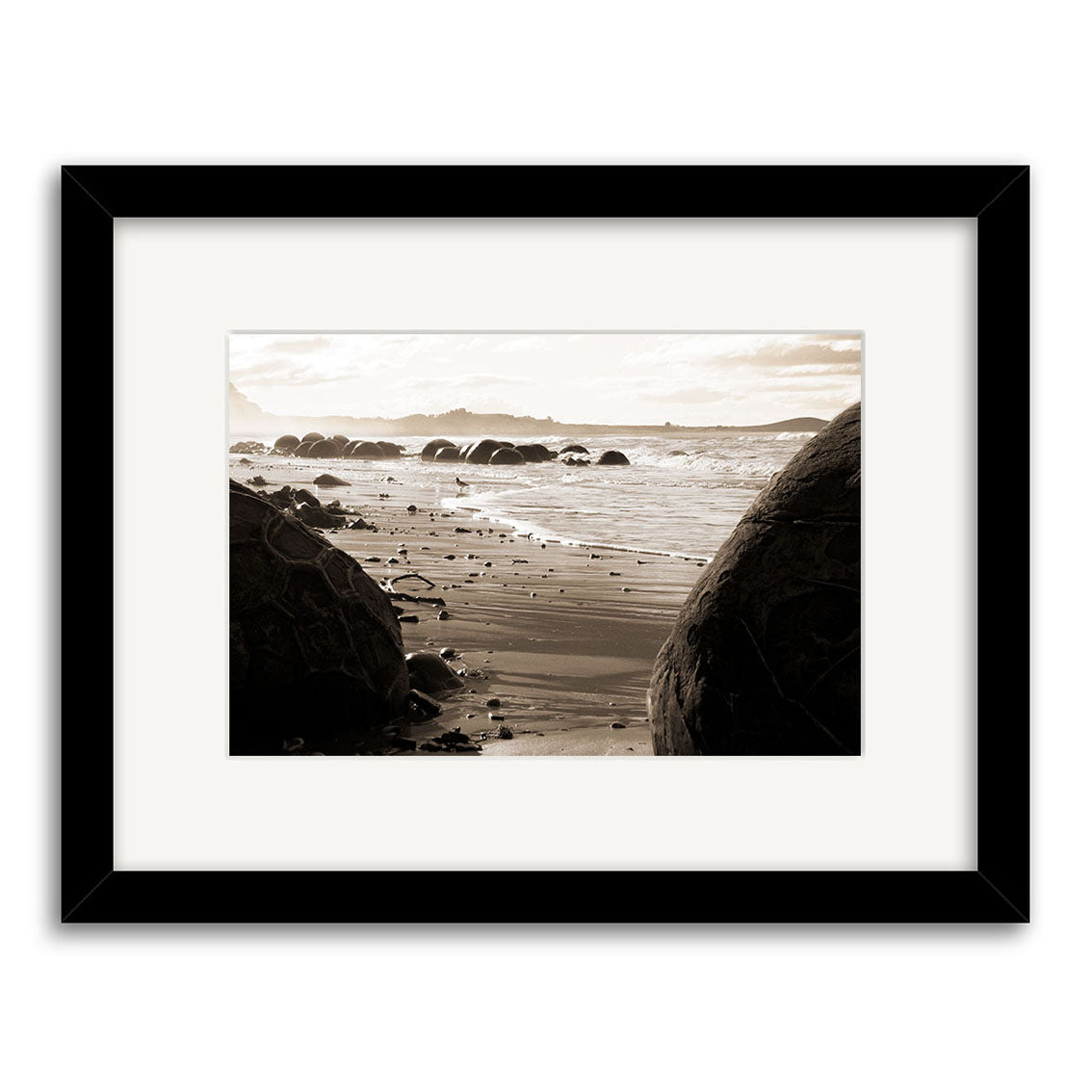 The Moeraki Boulders - RP056 is a framed black-and-white Reuben Price photographic print featuring large round boulders on a beach, with waves and distant hills, printed on archival quality matte paper.