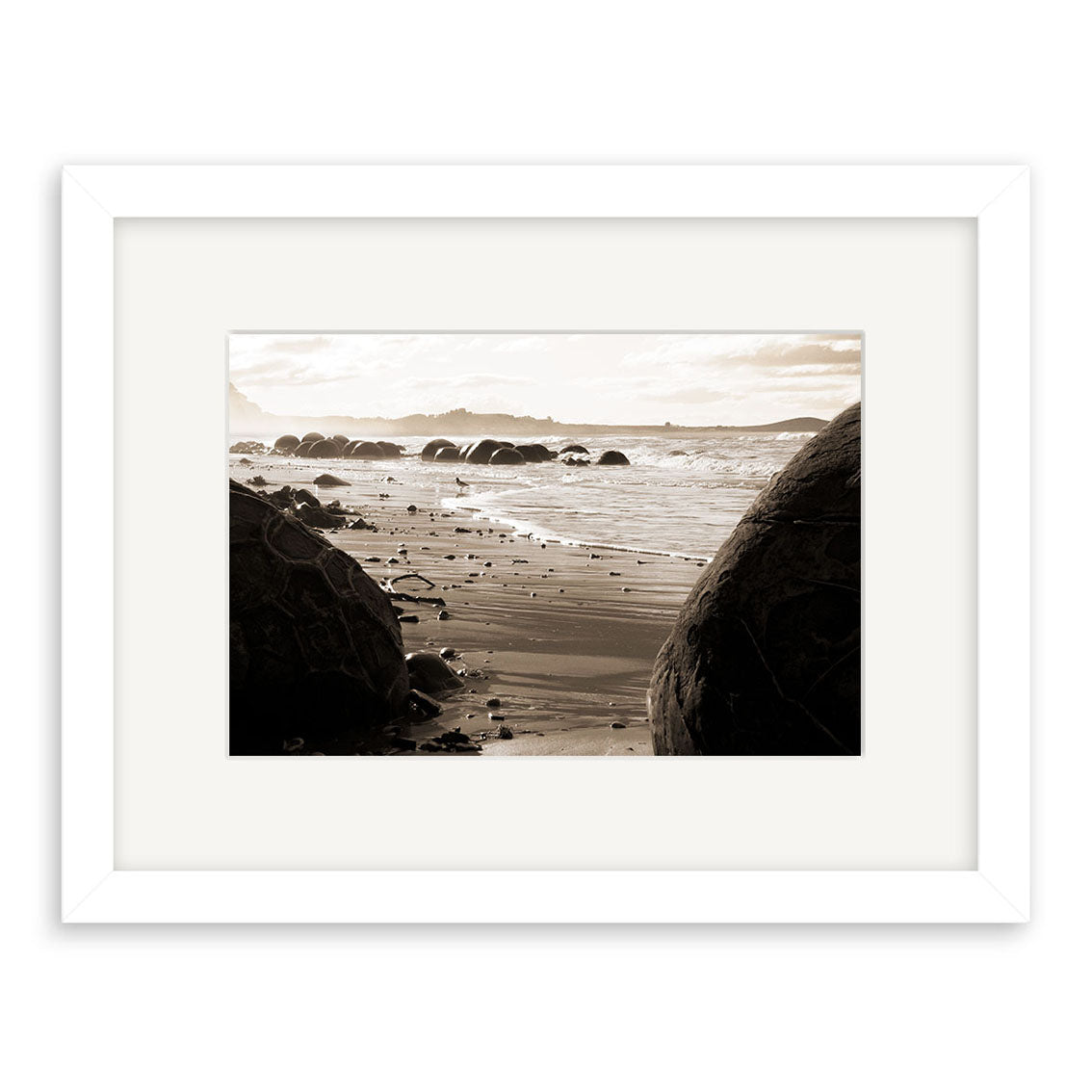 Moeraki Boulders - RP056: Sepia beach photograph featuring large round boulders on sand and in water, gentle waves, distant hills, and a cloudy sky. White-framed, archival matte paper—a classic New Zealand scene.