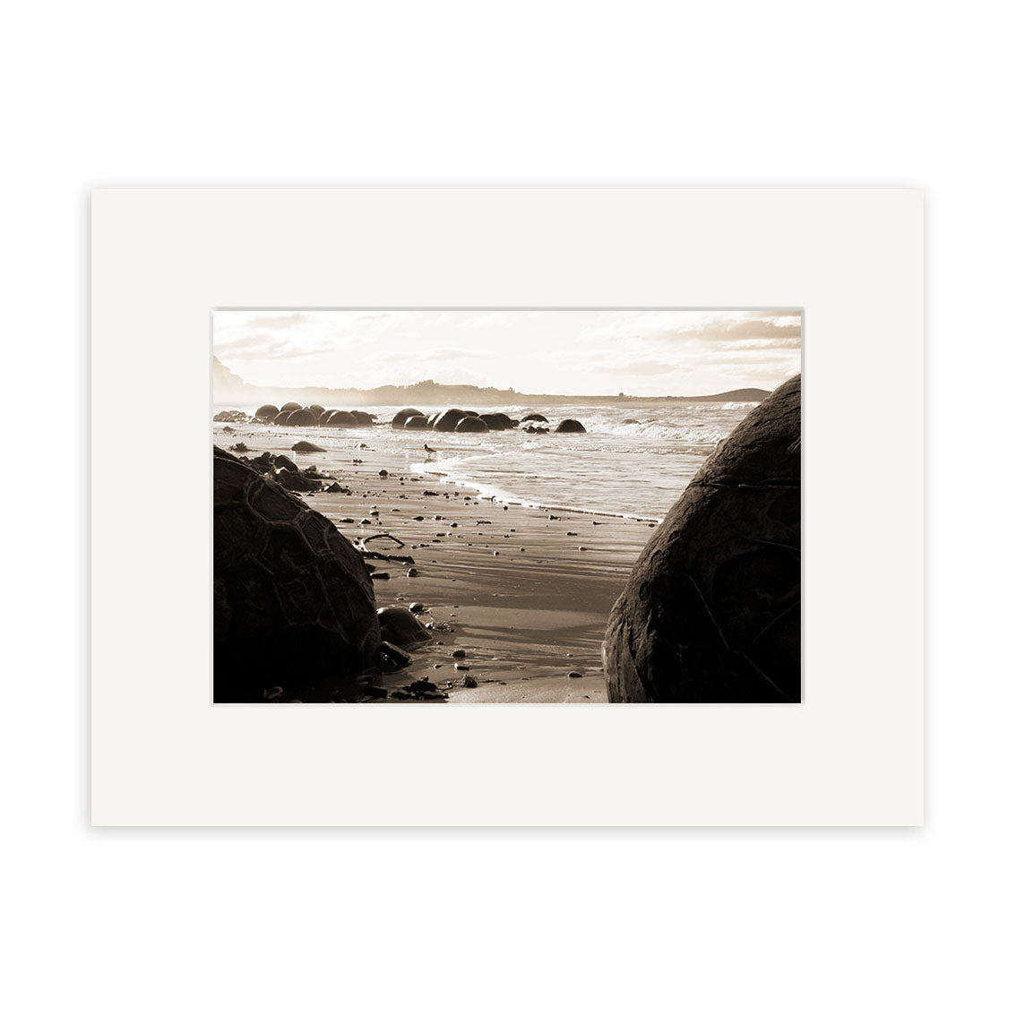 A sepia-toned photo of large, round boulders on a sandy beach with waves and mountains, Moeraki Boulders - RP056 is a Reuben Price photographic print on archival matte paper, presented in a white matte frame.