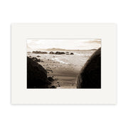 A sepia-toned photo of large, round boulders on a sandy beach with waves and mountains, Moeraki Boulders - RP056 is a Reuben Price photographic print on archival matte paper, presented in a white matte frame.