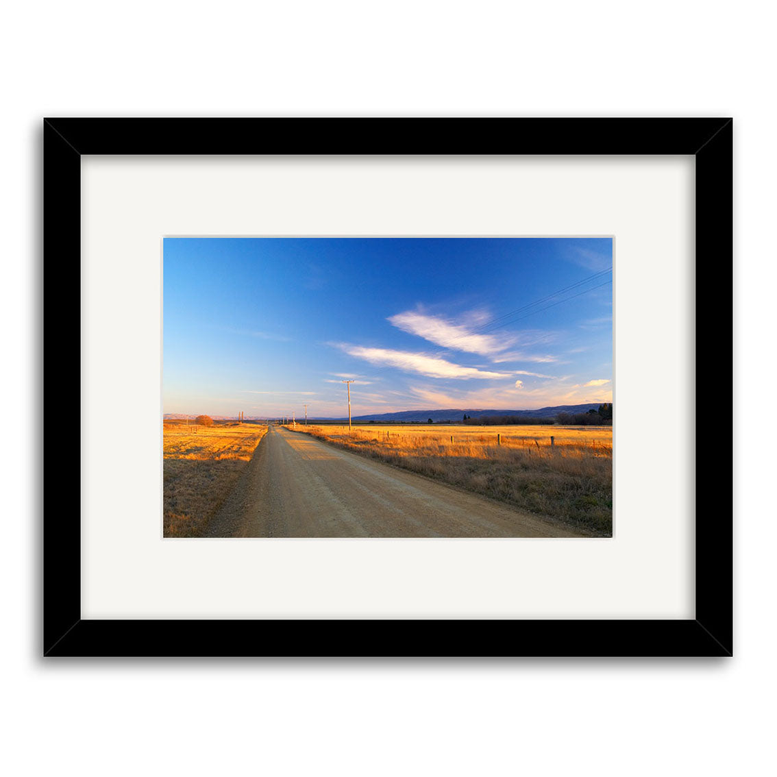 Wedderburn - RP057 by Reuben Price captures a rural dirt road under a blue sky, lined by fields and power lines. This New Zealand photographic print is printed on archival matte paper with a white matted border.