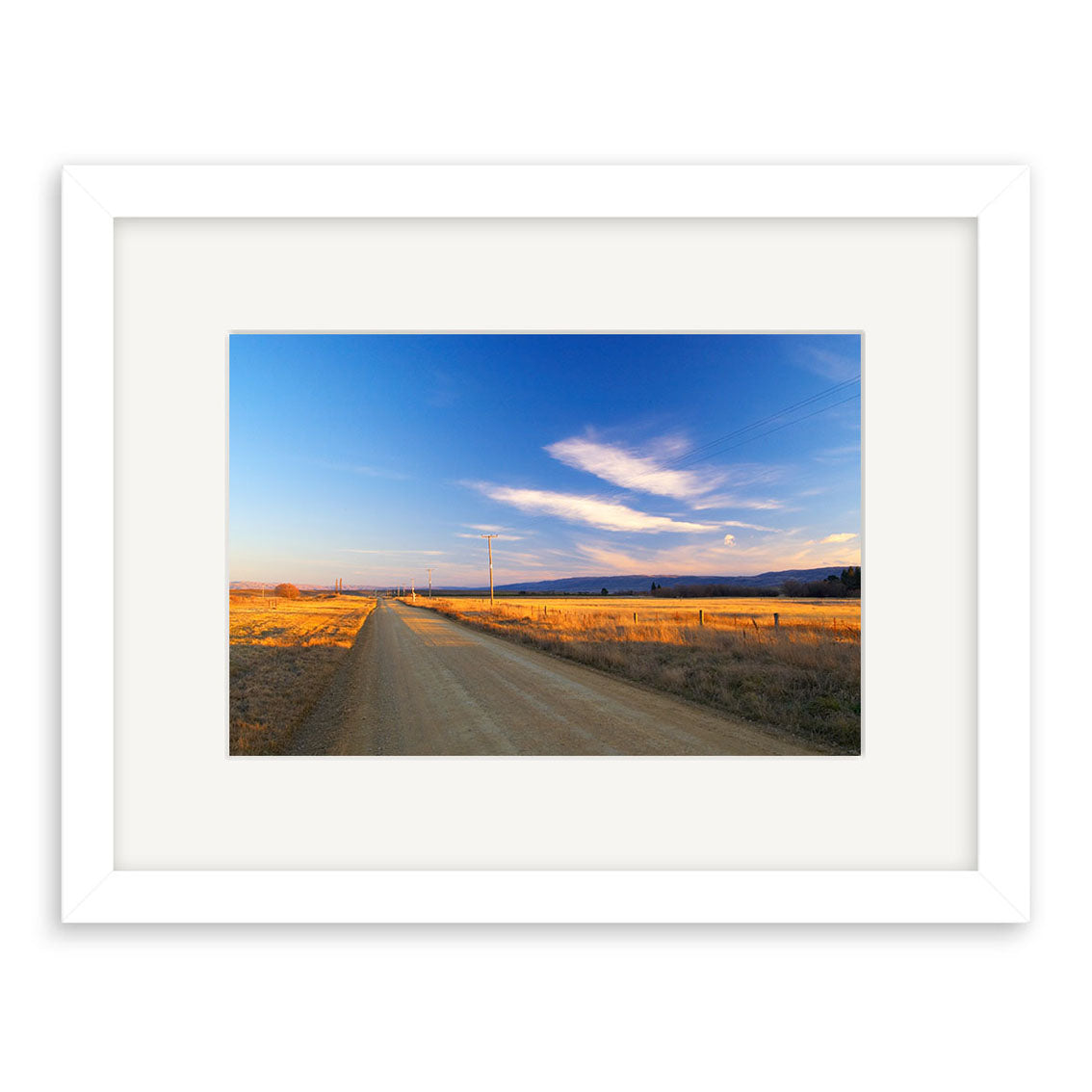 “Wedderburn - RP057” by Reuben Price is a framed New Zealand photographic print of a rural dirt road with power lines and golden fields under blue sky, printed on archival matte paper in a white frame with white mat.
