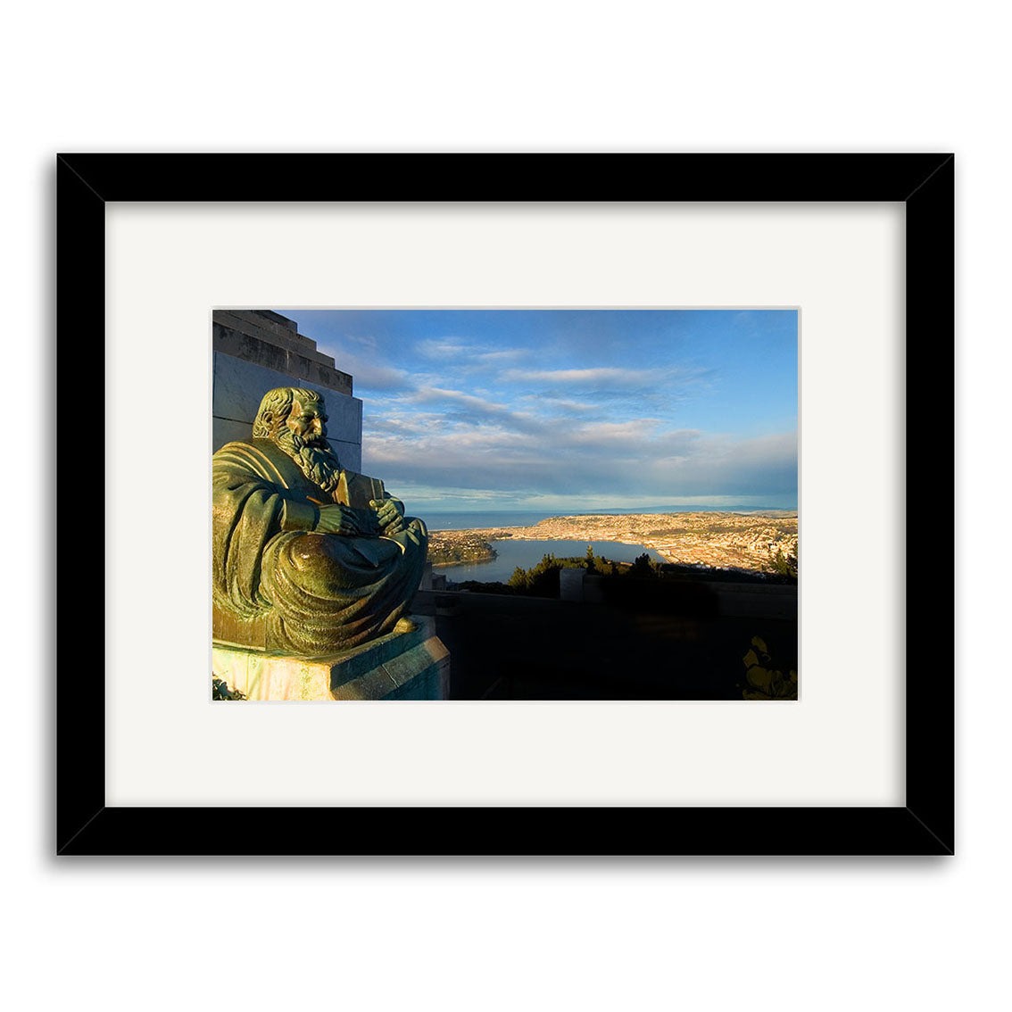 The photographic print Dunedin - RP059 shows a bronze seated statue in the foreground, a coastal New Zealand city and water in the background, all beneath a blue sky. It comes framed with a wide white mat and black frame.