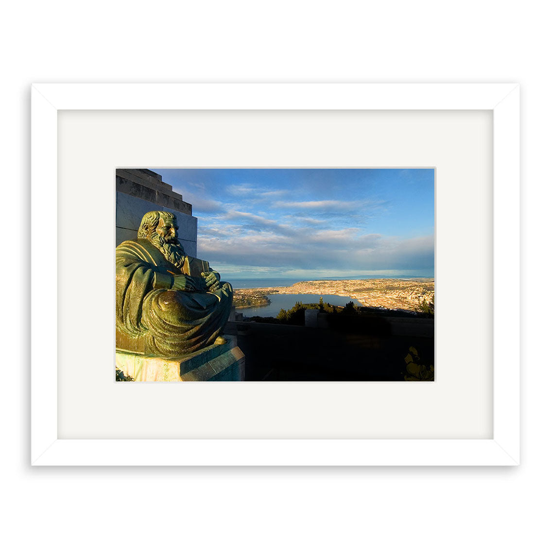 Dunedin - RP059 features a bronze statue of a seated figure overlooking coastal Dunedin, New Zealand, with city buildings and water under a partly cloudy sky. Available as archival quality matte paper photographic prints in a white frame.