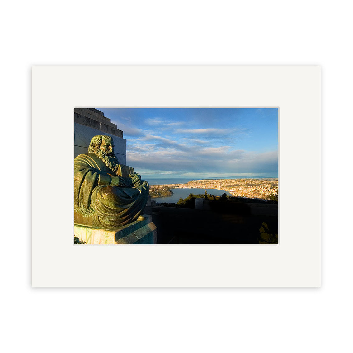 The Dunedin - RP059 print showcases a bronze seated statue overlooking the cityscape of Dunedin, NZ, with distant land and water beneath a blue sky. Presented with a white mat, its ideal for archival quality matte paper prints.