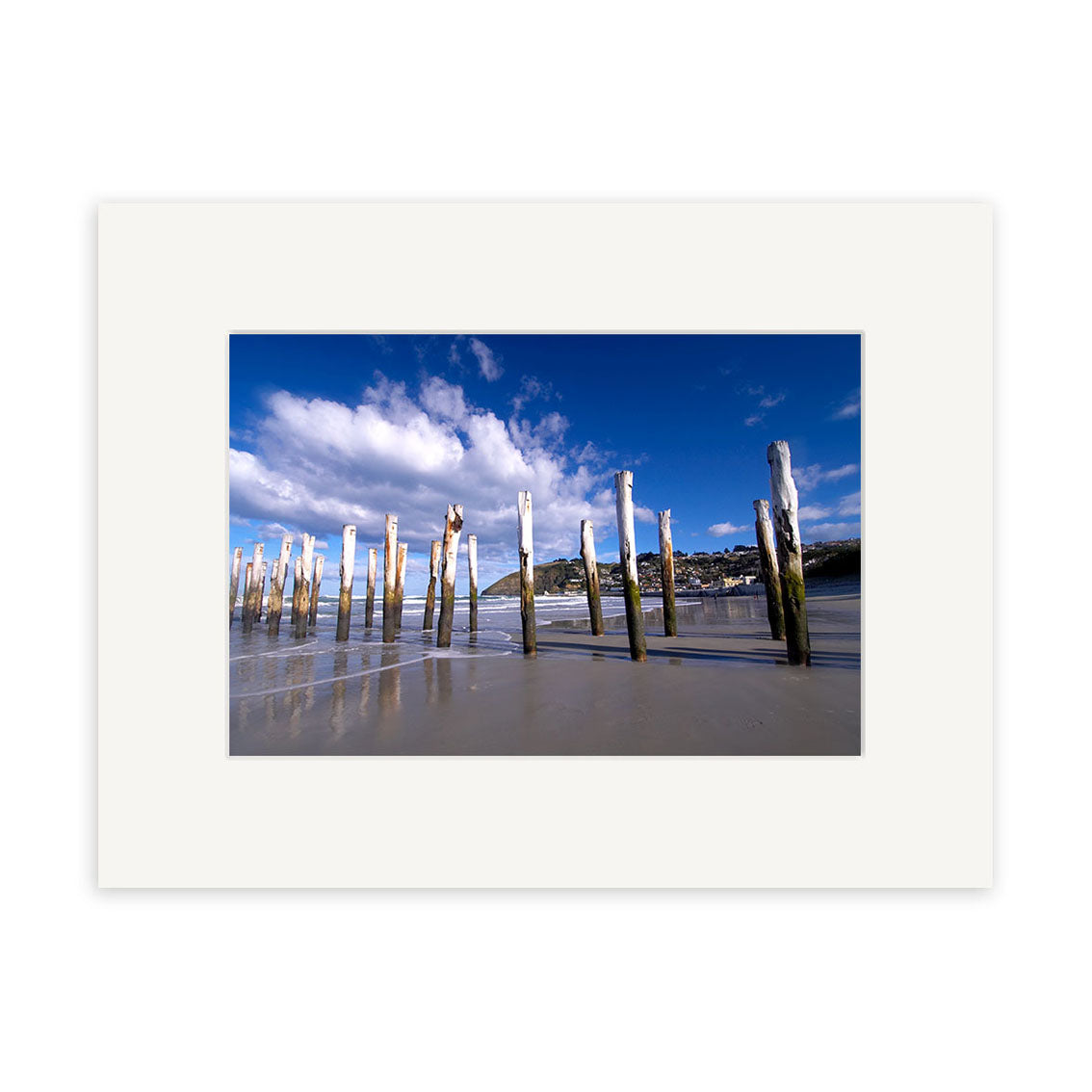 The photographic print St Clair - Dunedin - RP060 captures weathered wooden posts on a sandy beach under a blue sky, with calm water reflecting the posts and distant buildings. Available on archival quality matte paper.