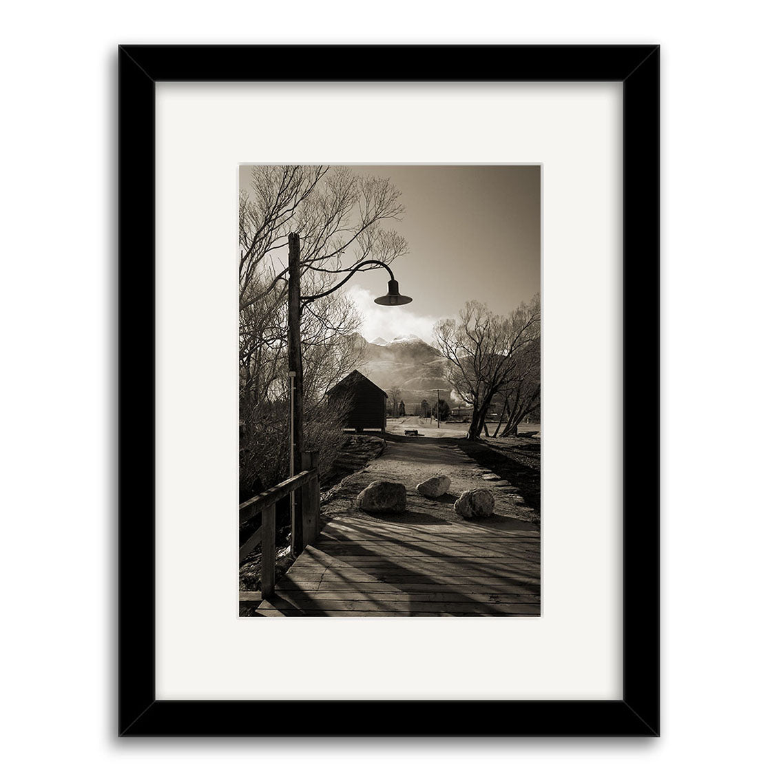 Glenorchy - RP062, a black-and-white photo by Reuben Price, features a rural New Zealand scene—wooden bridge, overhead lamp, hay bales, leafless trees, barn, and cloud-covered mountains—available as prints with a black border.
