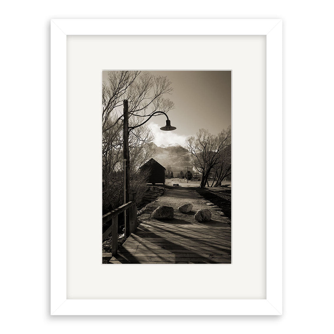 The black-and-white photo Glenorchy - RP062 features a white frame, a wooden bridge to a barn with mountains behind, bare trees, and a vintage street lamp along the path under a partly cloudy sky—evoking classic New Zealand scenery.