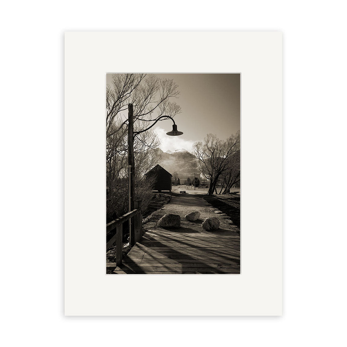 A sepia-toned photo titled Glenorchy - RP062 features a wooden bridge, two large rocks, a streetlamp overhead, bare trees, and a barn-like building with distant mountains—showcasing the timeless beauty of New Zealand by Reuben Price.