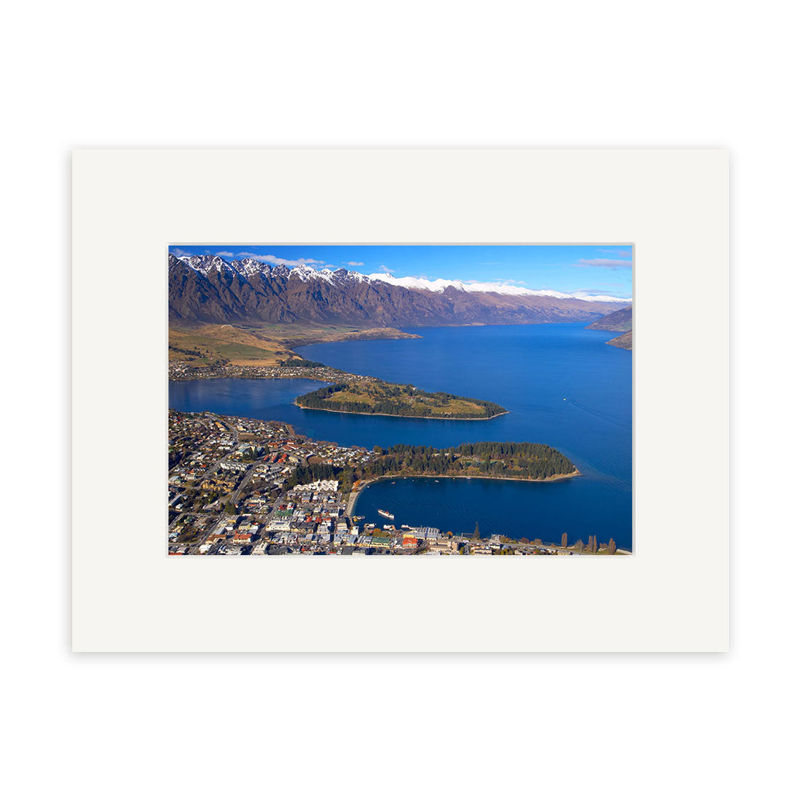 Aerial view of Queenstown (RP063), a New Zealand lakeside town with a peninsula and forested island, blue water, and snowy peaks under a clear sky. Available as photographic prints on archival quality matte paper.