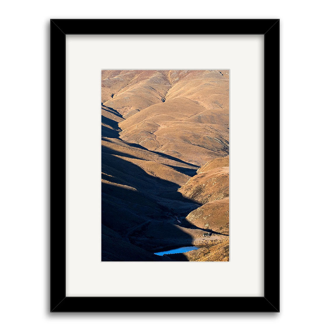 The Cardrona Valley - RP070 features a stunning landscape photograph of rolling brown hills with shadows, and a small blue pond near the bottom. Printed on archival quality paper, it comes in a black frame with white mat for an elegant display.