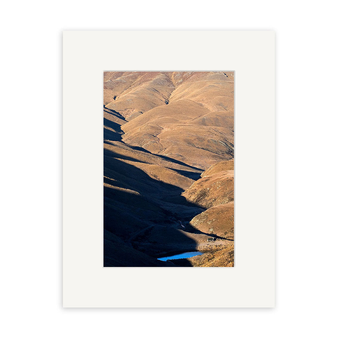 Cardrona Valley - RP070: A photo of rolling dry brown hills with sharp shadows and a blue water patch, printed on archival paper inspired by New Zealand landscapes, surrounded by a plain white mat board.