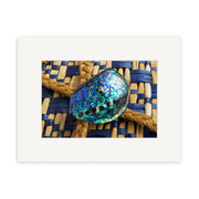 A close-up of the Paua Shell - RP085, iridescent and resting on a tan and navy woven surface, is showcased within a white mat—ideal for photographic prints on archival quality matte paper.