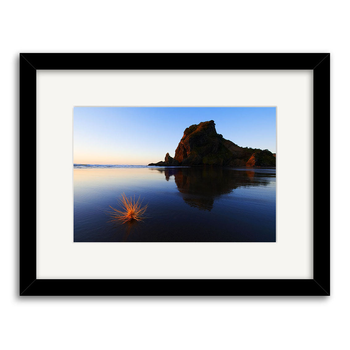 The Piha Sunset - Lion Rock - RP087 framed Reuben Price photography print features a coastal sunset with Lion Rock reflected in calm water and a small grass clump on wet sand, crafted as an exquisite archival quality print.