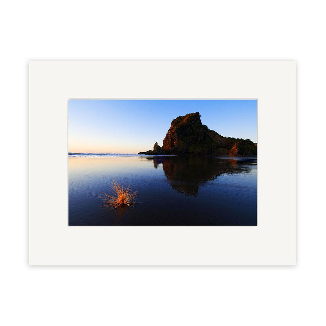 A large rock formation reflects in calm sunset water at Piha. Grasses stand out on smooth, wet sand as the sky shifts from yellow to blue—an exceptional New Zealand photographic print: Piha Sunset - Lion Rock - RP087 by Reuben Price.