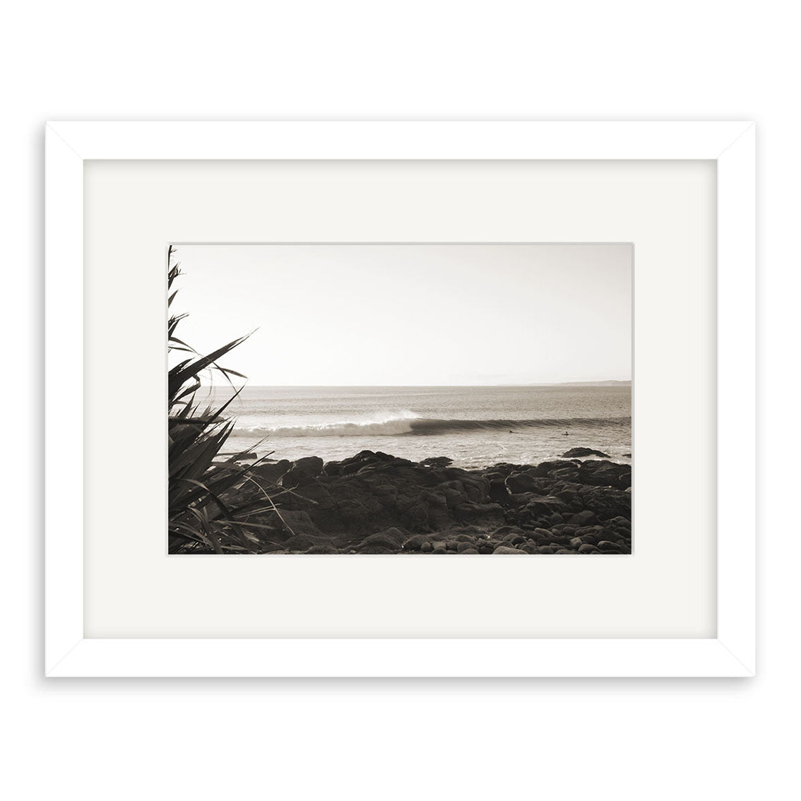 Whale Bay - RP091 is a framed black-and-white photograph of ocean waves crashing against a rocky New Zealand shore, with plants in the foreground. Printed on archival quality paper, its ideal for photography collectors.