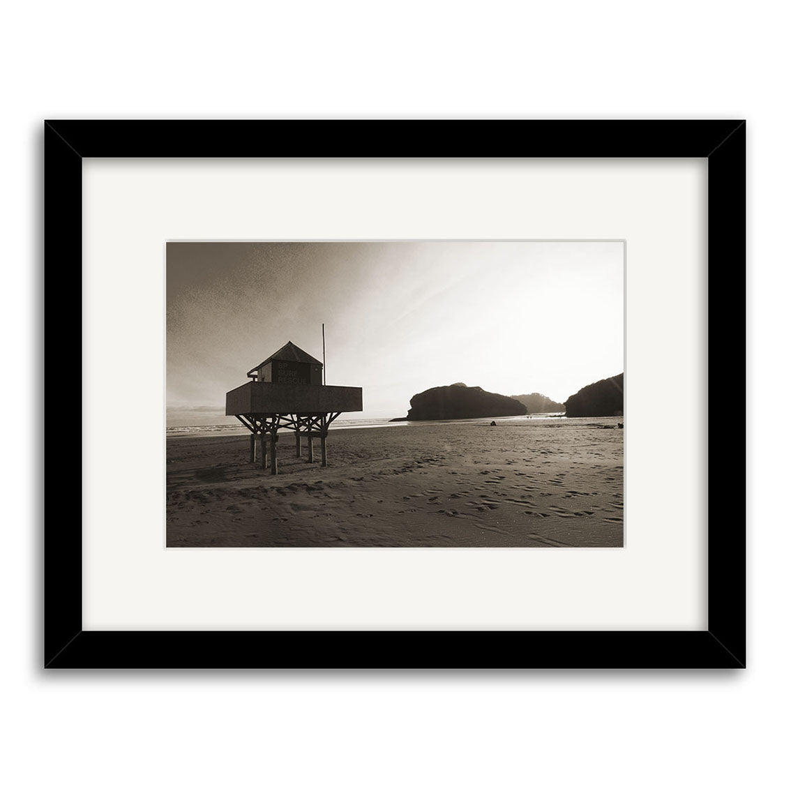 Bethells Beach - RP093: A black-and-white photo of a wooden stilt house on a deserted New Zealand beach with rocky cliffs, long sun-cast shadows, and framed with a white mat and black border.