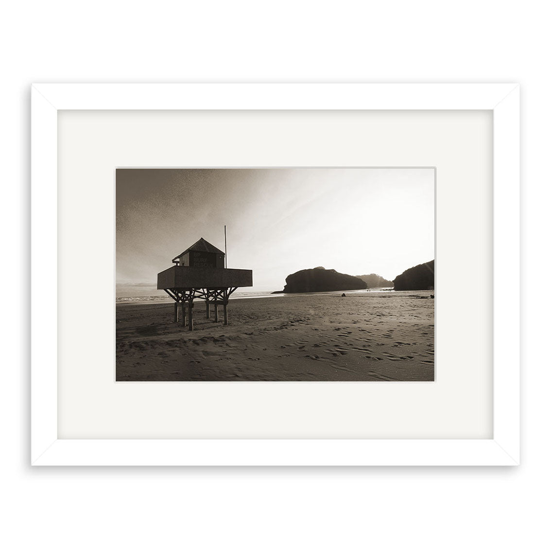 Bethells Beach - RP093: Black-and-white photograph of a lifeguard hut on stilts atop sandy New Zealand shores, rocky cliffs behind, long shadows from the setting sun. Photographic print displayed in a white frame.
