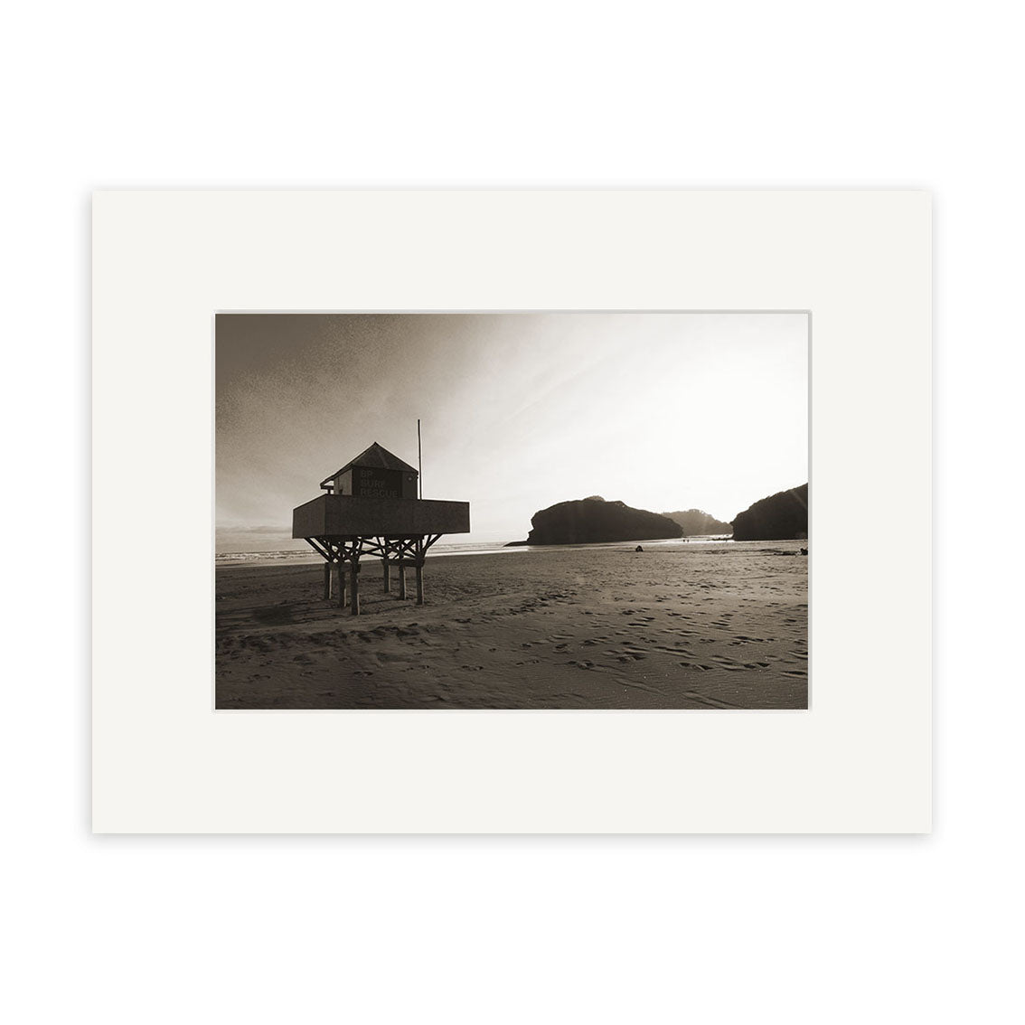 Bethells Beach - RP093: Black-and-white photo of a small wooden stilt building on a sandy New Zealand beach with rocky hills, low sun and long shadows. Available as photographic prints on archival quality matte paper.