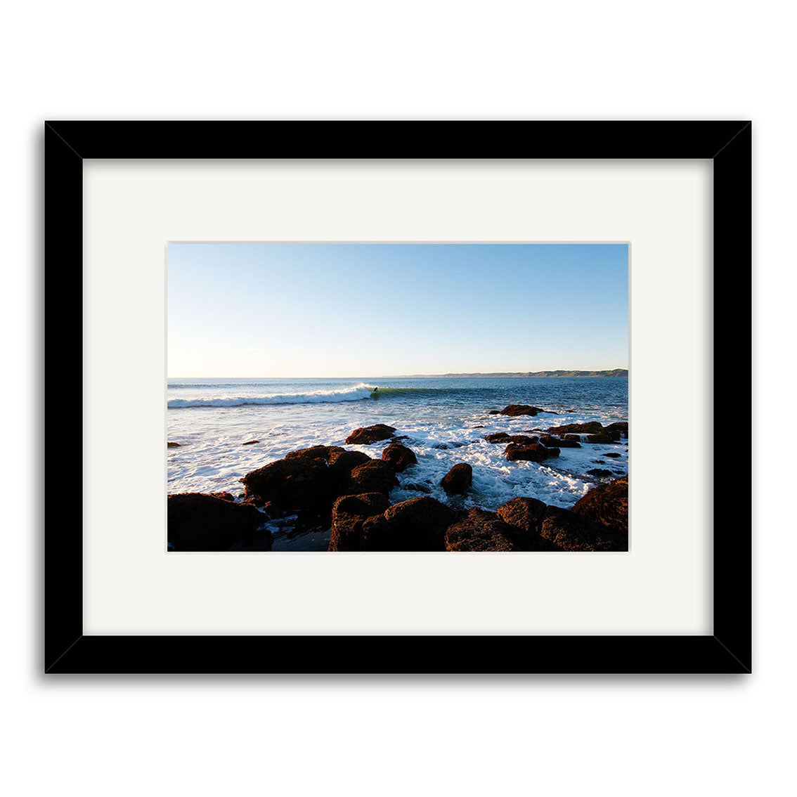 Manu Bay - RP094: A framed photo of a New Zealand rocky shoreline with waves under a blue sky, centered in a white mat and black frame. Printed on archival quality matte paper.