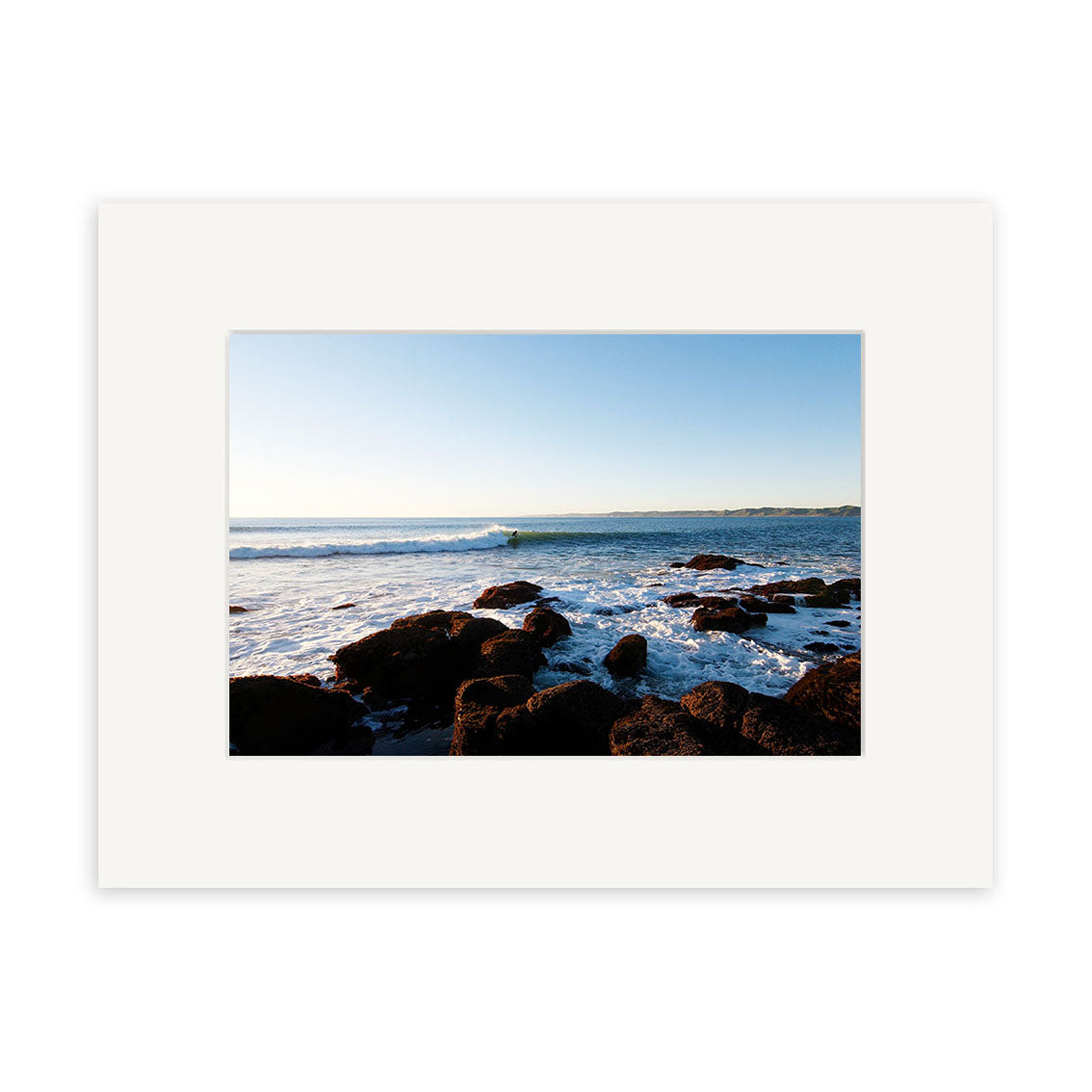 A photo titled Manu Bay - RP094 captures a rocky New Zealand shoreline with waves beneath a clear blue sky, presented in a white mat board for displaying as archival quality matte paper prints.