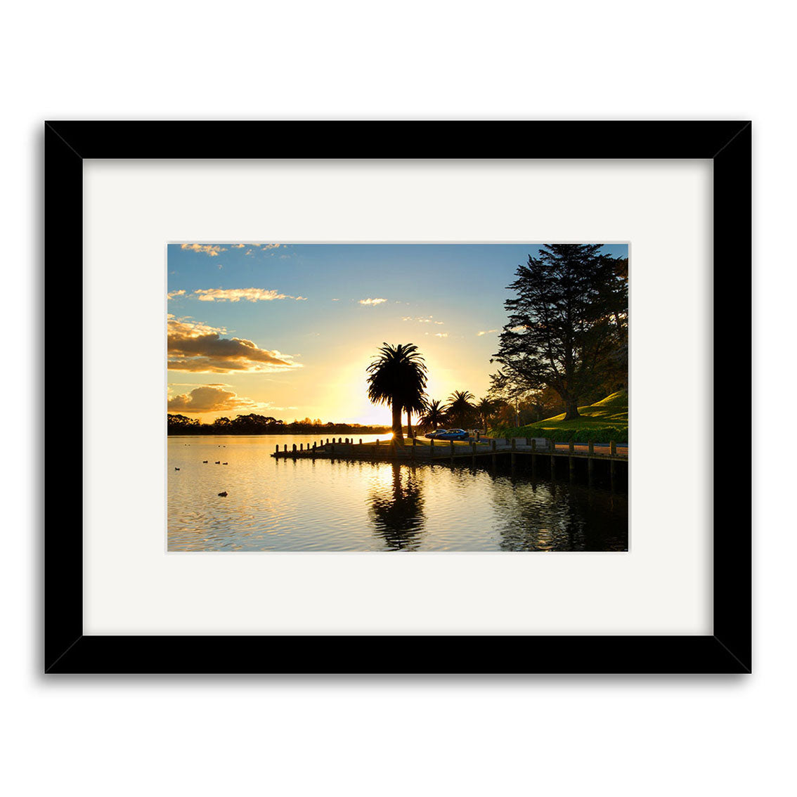Hamilton Lake - RP097: A framed archival matte print featuring a sunset over New Zealands calm lake, silhouetted palm trees, wooden dock, and grassy shoreline beneath a clear sky with light clouds.