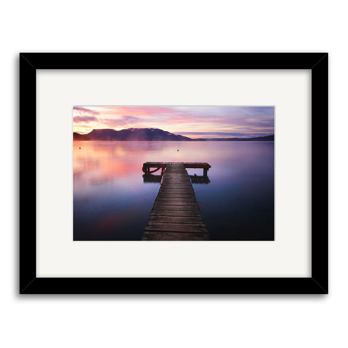 A wooden dock stretches into a calm lake at sunset with mountains and vibrant skies behind. Lake Tarawera - RP098 by Reuben Price is an archival quality print, elegantly framed with a white mat and black frame.